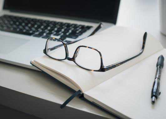 Un ordinateur portable, une paire de lunettes, un cahier ouvert et un stylo.
